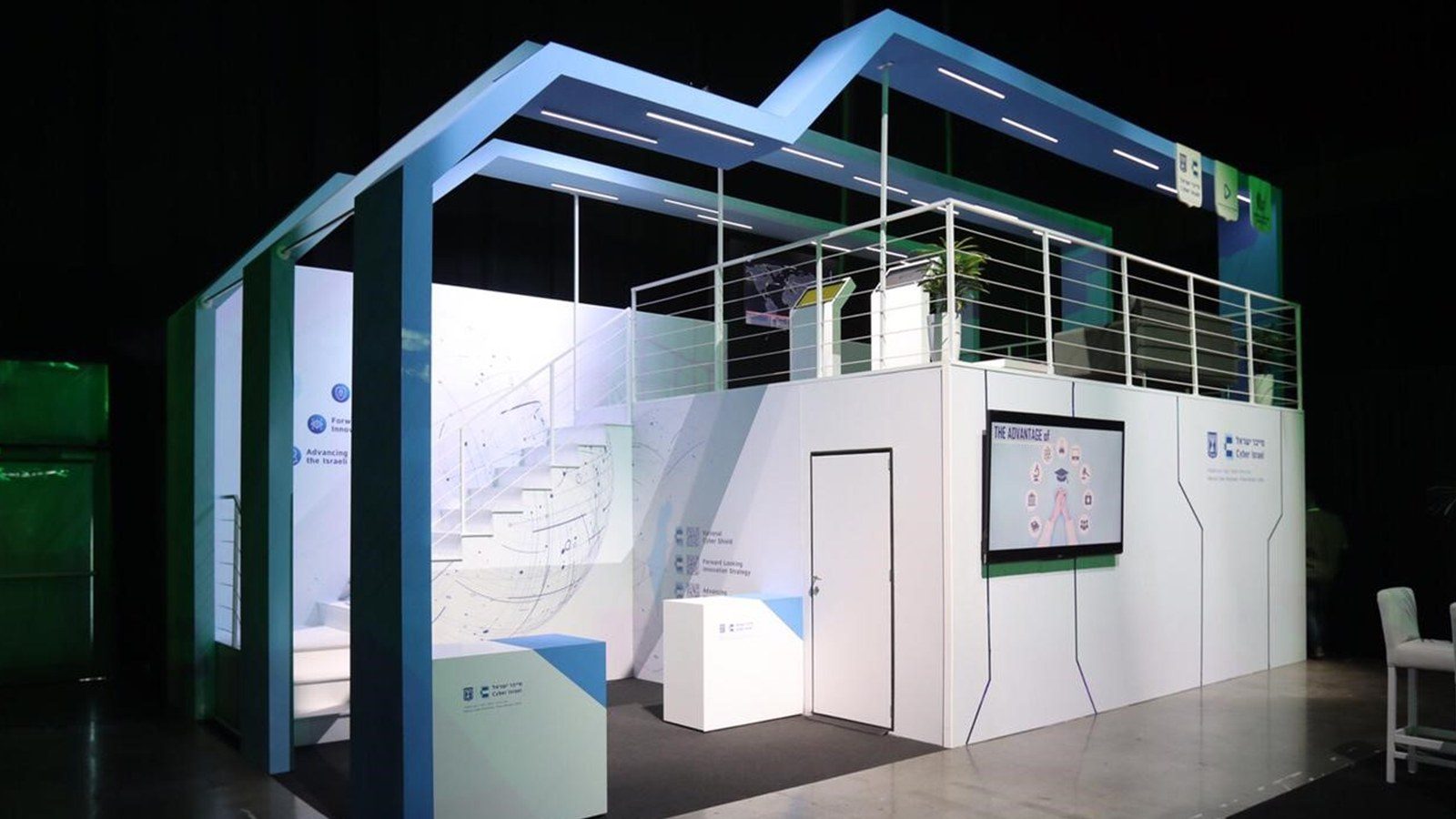 A two-storey exhibition build standing in a darkened hall, an open upper deck with railing above a white lower floor.