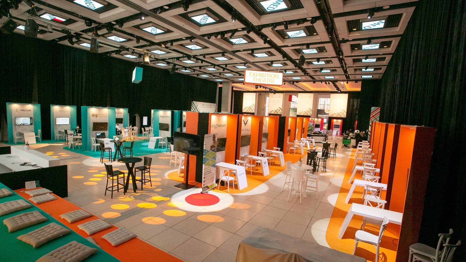 An exhibition floor of built bays in teal and orange, an illuminated Exhibition Theatre sign overhead.