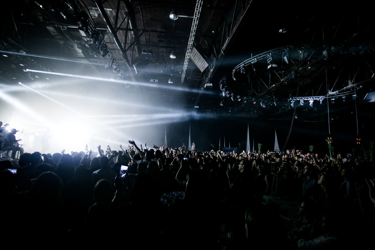 A full hall at the moment of the show: beams cutting through haze from the stage, lighting rigged on trusses overhead and the crowd with their hands up.