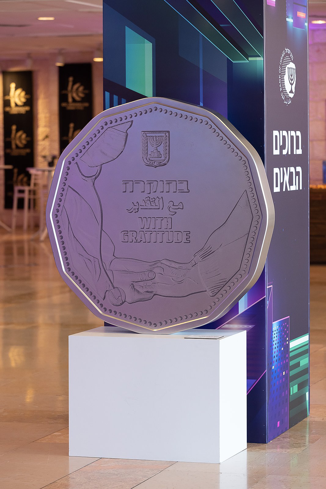 A lobby installation: an oversized replica of a coin on a white plinth, its face inscribed WITH GRATITUDE in Hebrew, Arabic and English, beside a branded welcome totem.