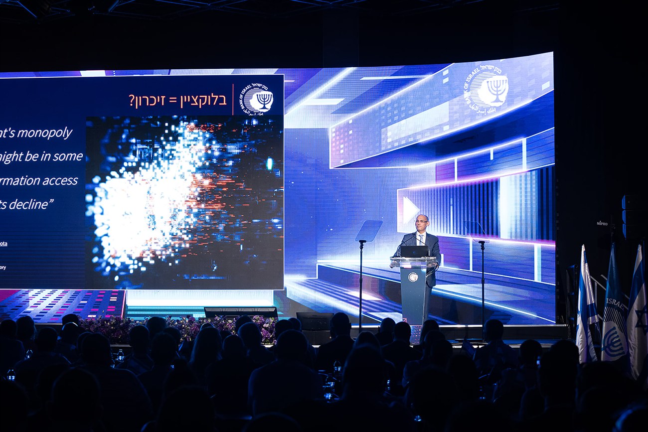 A speaker at the lectern against a stage set built as a full-width LED wall, a presentation slide on the screen to one side and the audience in silhouette.