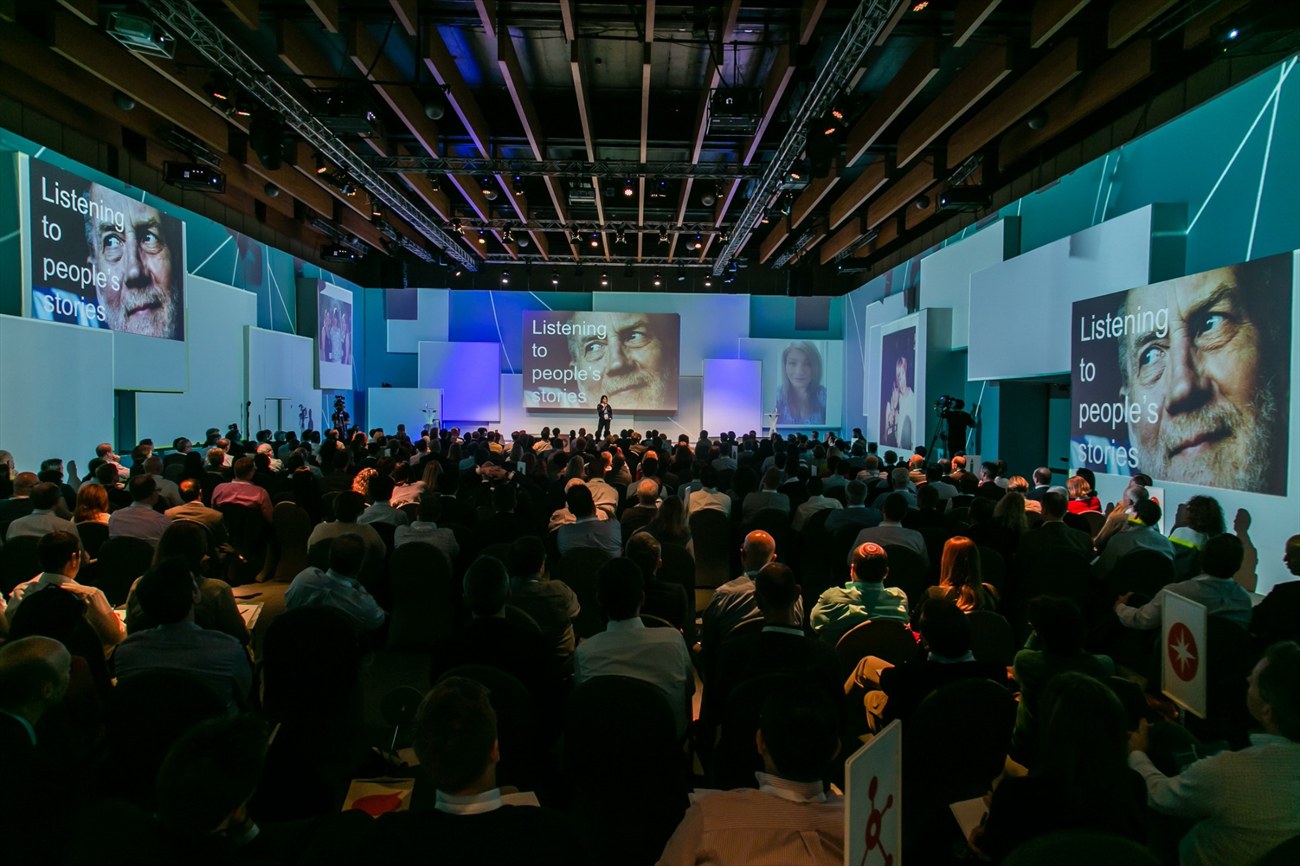 A full plenary facing a stage backed by a full-height wall of portraits.