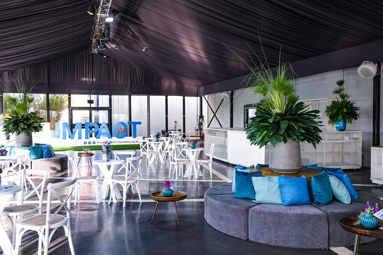 The lounge pavilion in daylight before the doors open, white cafe furniture under a draped roof and the conference mark standing on the lawn beyond the glass.