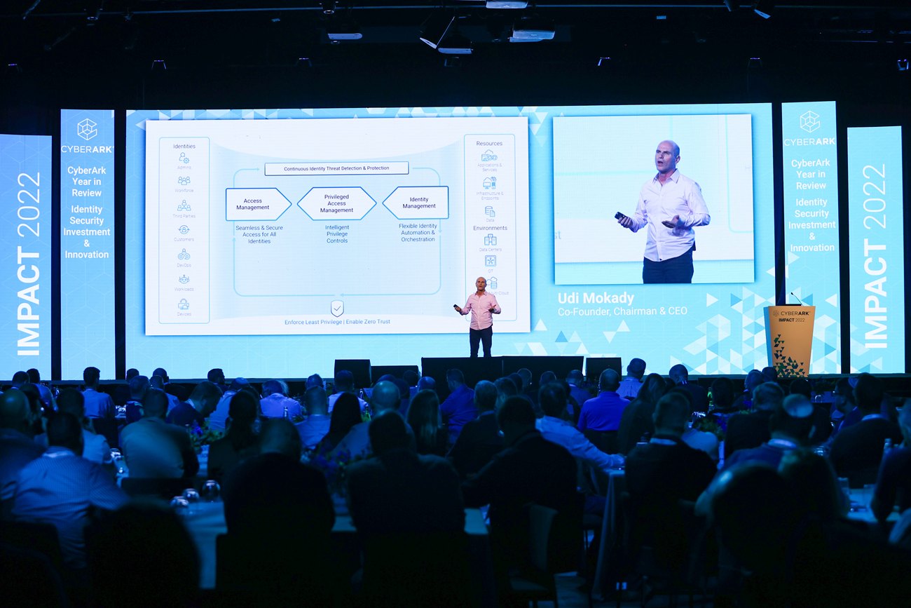 The main stage seen over the audience: a full width screen, the conference mark on the flanking panels and the hall seated in front of it.
