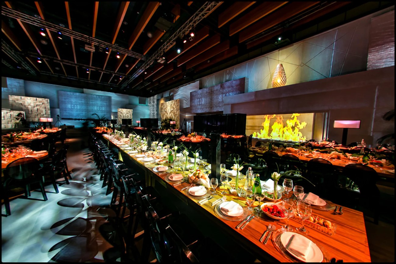 A long banqueting table running the length of a room dressed for dinner, a wall of open flame beside it.