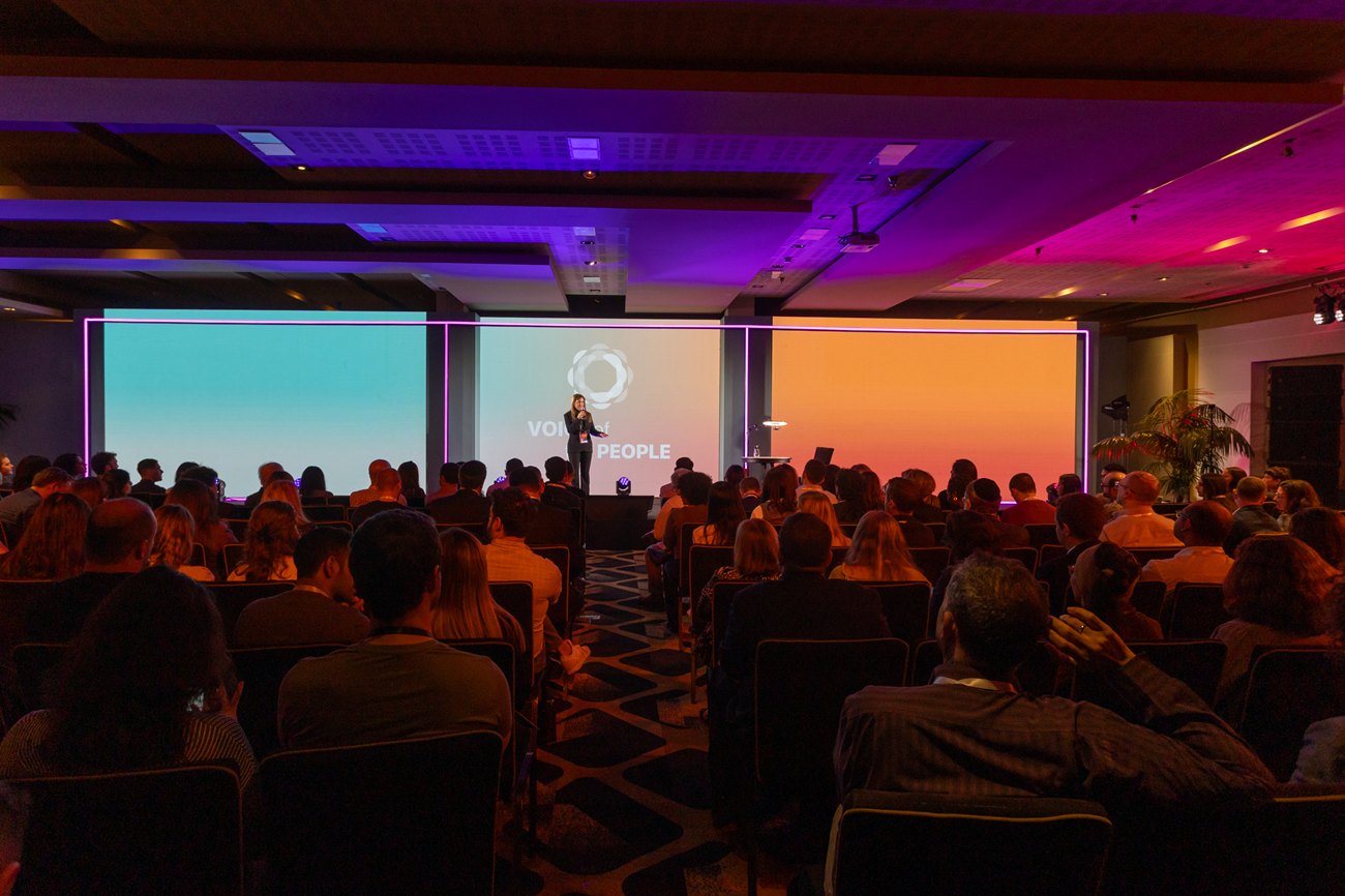 The hall in session, a speaker on stage between three lit screens and the audience seated in front of them.
