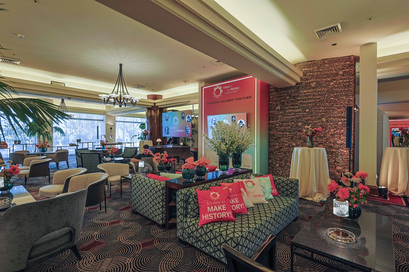 The hospitality floor seen wide: low seating and branded tables across the room, a lit totem against the stone wall and daylight through the windows.