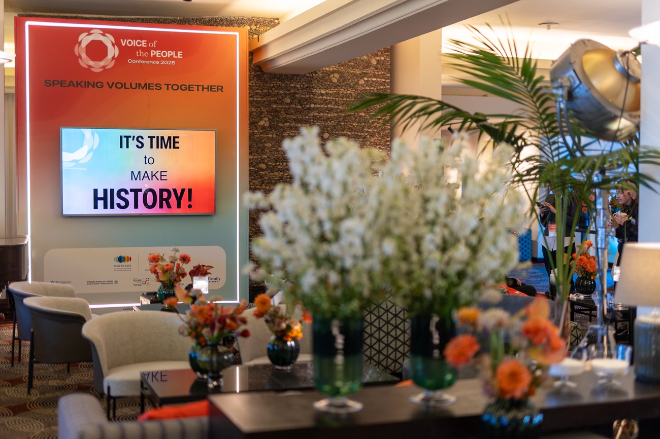 A hospitality corner: a branded panel with a screen behind a tall arrangement of white and orange flowers.