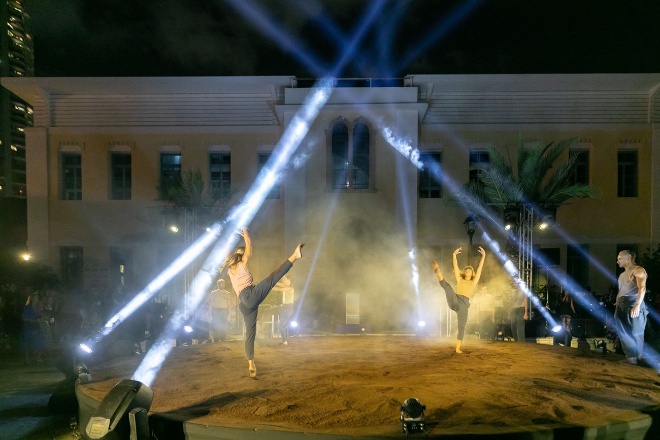 Dancers on a round sand stage at night, beams of light crossing above them in front of a historic building.